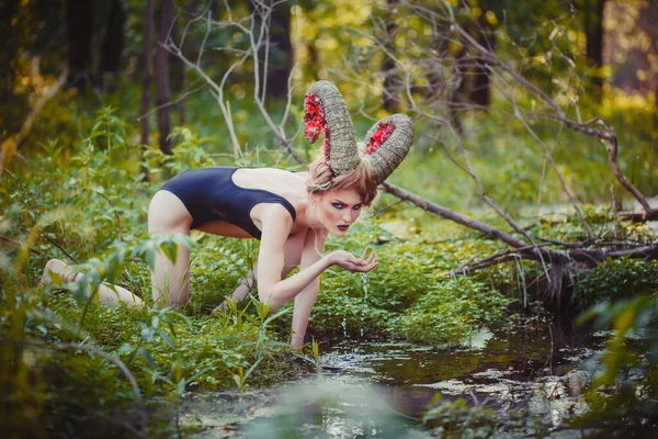 Sexy beautiful woman with huge horns drinks water from the river like an animal. Fantasy girl in the forest