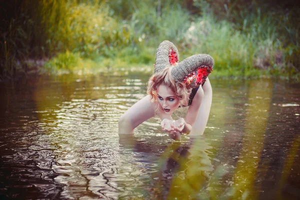 Sexy beautiful woman with huge horns drinks water from the river like an animal. Fantasy girl in the forest