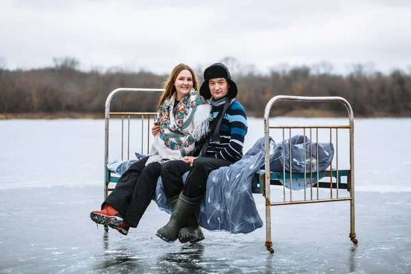 A couple in love is sitting on an iron bed and embracing. Cold, frozen lake, snow all around