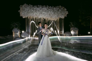 Damat, düğün kışı süslemelerinin arka planında boyalı kanatları olan gelini kucaklıyor. Dondurucu ve ışıklandırma ile gece fotoğrafçılığı teknikleri.