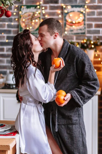 People in dressing gowns kiss in the kitchen, decorated for Christmas. They have oranges in their hands