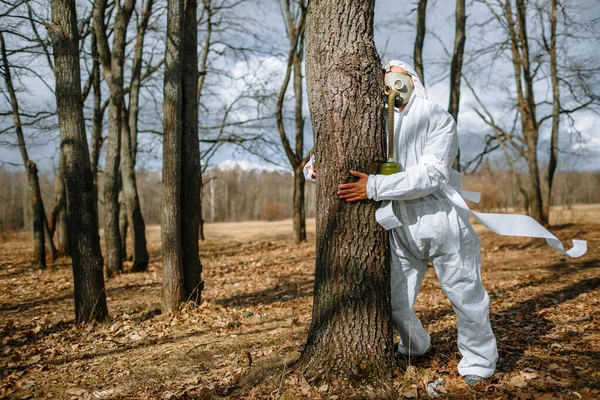 The man in the medical suit and a gas mask wrapped around a tree in the forest. The suit is tied with toilet paper