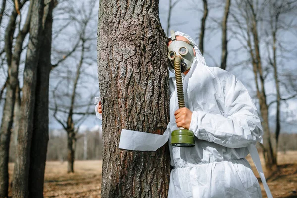 The man in the medical suit and a gas mask wrapped around a tree in the forest. The suit is tied with toilet paper