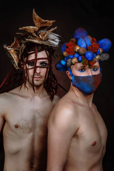 Two male models. A model with an egg - shaped headdress of cones and a mask on her face. Model with a headdress made of tree bark and ropes