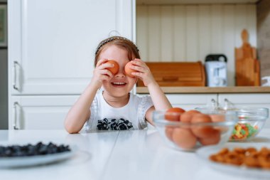 Gülümseyen bir kız, kek yapmak için malzemelerin bulunduğu mutfak masasında oturuyor. Kız gözlerini tavuk yumurtasıyla kapadı.