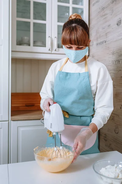A female cook in a medical mask and gloves whips up cake batter