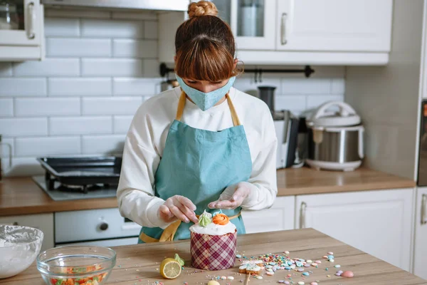 A cook in a medical mask and gloves lays out sweet figures on the icing of an Easter cake