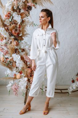 Açık beyaz bir yaz elbisesi içindeki model kız bir çiçek kemerinin arka planında. Galeri için kıyafet gösterisi.