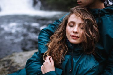 Yeşil yağmurluklu sevgililerin portresi, bir taşın üzerinde oturuyor, bir şelalenin arka planında