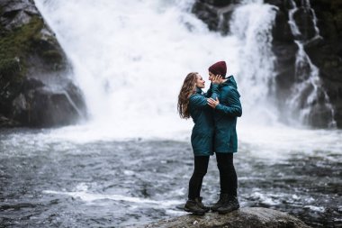 Yeşil yağmurluklu sevgililerin portresi, bir taşın üzerinde duruyor, bir şelalenin arka planında.