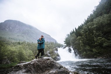 Yeşil yağmurluklu sevgililerin portresi, bir taşın üzerinde duruyor, bir şelalenin arka planında.