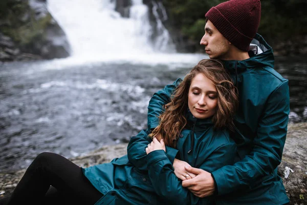 滝を背景に岩の上に座っている緑のレインコートの恋人たち — ストック写真