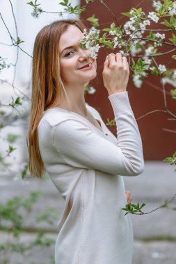Beyaz bluzlu gülümseyen bir kız kiraz çiçeğinin içinde duruyor.