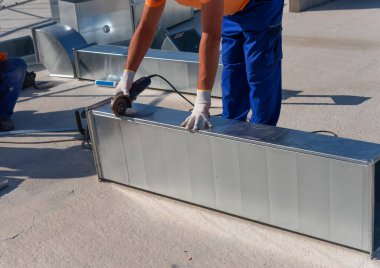 HVAC technician is working on a roof of new industrial building. Close-up view of the young technician repairing an air duct with the angle grinder. HVAC worker cutting ductwork with an angle grinder. clipart
