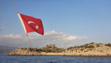  Türkiye 'nin kırmızı bayrağı adanın üzerinde mavi gökyüzüne karşı. Önde mavi gökyüzüne karşı yatın halatında Türkiye 'nin kırmızı bayrağı var. Arka planda mavi denizde sarı kayalık bir ada var..
