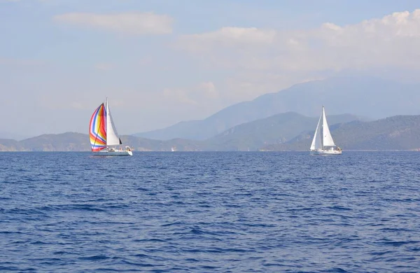 Two white sailing yachts sail one after another from right to left.  Blue sea. A yacht with a multi-colored sail leads the race. In the background on the horizon are several rows of mountains. Blue sky with clouds. Shot in the Mediterranean Sea.