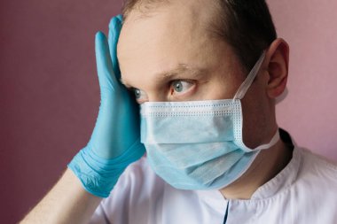 Düşünen doktor. Üzgün ve gergin doktor. Mavi cerrahi maskeli ve eldivenli bir doktorun yakın plan portresi. Elleri başının üstüne koyulmuş. Pandemik koronavirüs. Karantinaya alın. Virüs konsepti. 