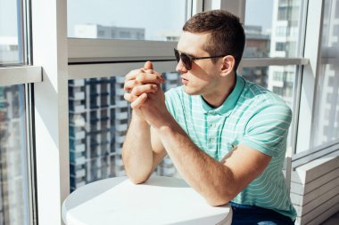 Mavi tişörtlü ve güneş gözlüklü bir adam masaya oturur ve pencereden dışarı bakar.. 