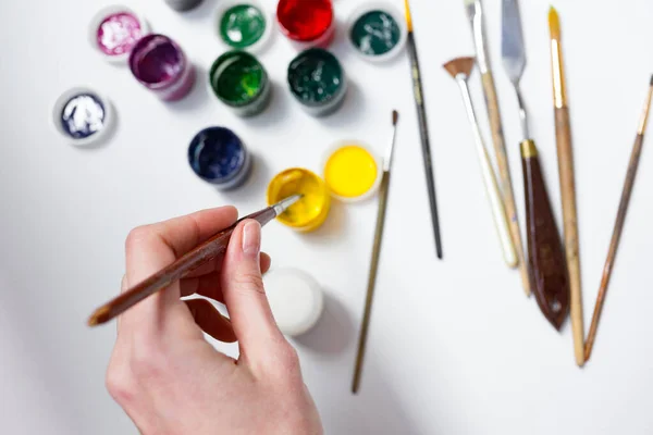 Akrylfärger, borstar och palettkniv på den vita duken. En process för målning. En hand med pensel tar en gul färg. Hobby. — Stockfoto