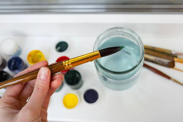 Farby akrylowe, szklany słoik, szczotki i nóż paletowy na parapecie. Ręka ze szczotką. Proces malowania. Włóczęga. — Zdjęcie stockowe