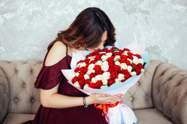 Kırmızı ve beyaz güllü büyük bir buketi olan güzel elbiseli bir kız evdeki kanepede oturuyor. Mutlu yıllar..