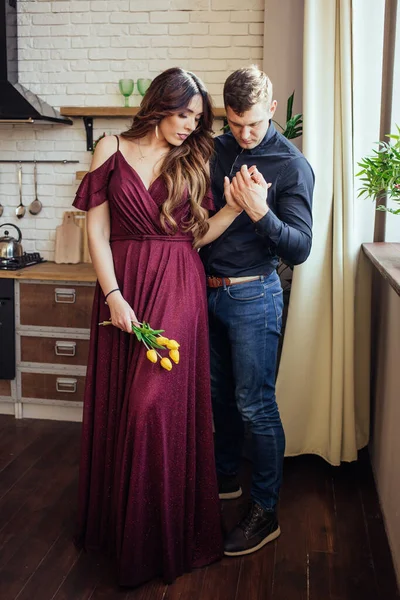一对可爱的夫妇站在厨房里.她的手在他手里一个穿着衣服，手里拿着黄色郁金香的女人。和谐的家庭浪漫的爱情故事幸福. — 图库照片