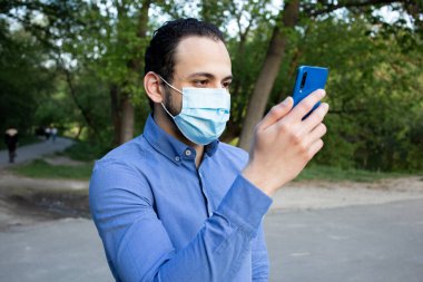 Ortadoğulu bir gencin parkta koruyucu maskeli portresi. Bir adam telefonuna bakar, video görüşmesiyle konuşur. Sosyal mesafe. 2020 pandemik koronavirüs. Karantinaya alın. Virüs konsepti.