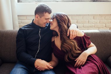 Aşıklar birbirlerine sevgiyle bakar. Evlenmek. Genç bir ailenin aşk hikayesi. Elbise giymiş gülümseyen bir kadın ve gömlekli bir adam kanepede sarılıyorlar. Ev rahatlığı. Yaşam biçimi.