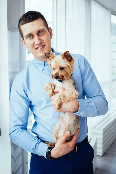 Un hombre feliz con una camisa azul sostiene a un perro en sus manos y sonríe en casa. Ventanas panorámicas en el fondo. Confort hogar . —  Fotos de Stock