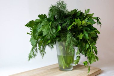 Cam bardakta taze dereotlu maydanoz ve beyaz arka plandaki kesme tahtasında su var. Baharatlı sebzeler masada. Sağlıklı vejetaryen yemekleri.