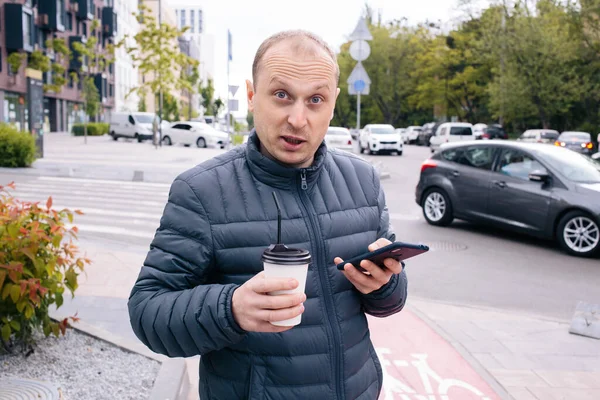Un caucasien positif surpris homme dans une veste grise avec une tasse en papier blanc de café Latte ou thé avec couvercle et paille et téléphone sur sa main sur le fond de la ville. Eco. Pause. Gros plan — Photo