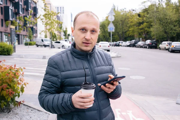 Un homme caucasien positif dans une veste grise avec une tasse en papier blanc de café Latte ou thé avec couvercle et paille et téléphone sur sa main sur le fond de la ville. Eco. Pause. Gros plan — Photo