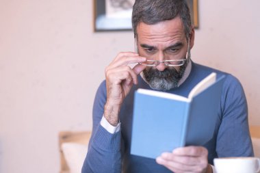 Gözlükleriyle kitap okumakta sorun yaşıyor olan yaşlı adam, kahve dükkanını çevreleyen