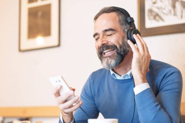 Kıdemli yaşlı adam büyük kulaklıklarla en sevdiği müziği dinliyor, müziğin zengin sesinin tadını çıkarıyor, bir kafede oturuyor.