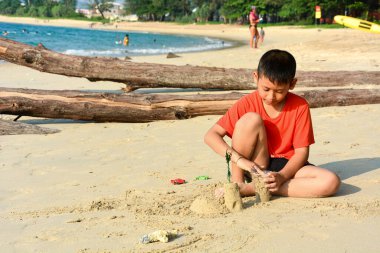 Genç Asyalı çocuk tropikal güzel sahilde kumdan bir kaleyi köpürtüyor. Akşamları güneş ışığı altında, Phuket Tayland.