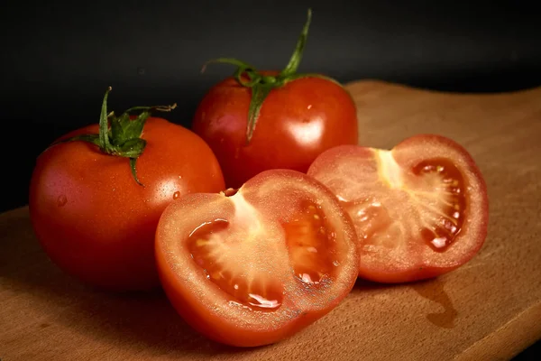 Rote Tomaten auf einem Holzschneidebrett, ganz und in Scheiben geschnitten auf schwarzem Hintergrund, mit leichtem Blu — Stockfoto