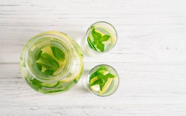 Işık ve taze salatalık, kavanozda limon ve nane içeceği ve beyaz masada bardaklar. Detoks suyu.