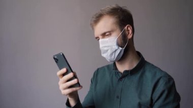 Sakallı ve maskeli bir adam telefonunda beyaz bir duvarın arkasına bakıyor.