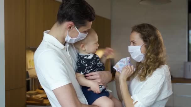 마스크를 쓴 어린 아버지와 어머니는 아이에게 마스크를 쓰려고 하지만, 아이는 마스크를 쓰는 것을 좋아하지 않는다. 세계적 유행병 기간에 격리 된 가정의 가족 — 비디오