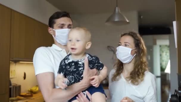 Молодые отец и мать в масках держат своего ребенка на руках, находясь дома в карантине во время пандемии — стоковое видео