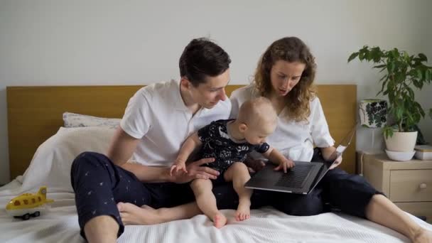 Los padres jóvenes con un niño se sientan en una cama con una computadora portátil, el niño intenta presionar botones en la computadora portátil — Vídeos de Stock