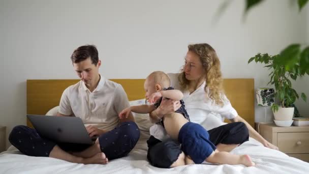 Os pais jovens e seu filho se sentam na cama, o pai tenta trabalhar em um laptop enquanto a criança o impede. Instrução — Vídeo de Stock