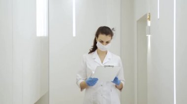 Maskeli bir kadın doktor koridorda yürüyor ve salgın sırasında yeni modern klinikteki hastalığın tıbbi kayıtlarını okuyor.