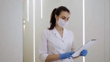 Beyaz cüppeli, maskeli ve eldivenli bir kadın doktor tıbbi kayıtları okuyor ve salgın sırasında yeni bir klinikteki kameraya teşhisi bildiriyor.