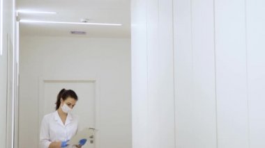 Maskeli bir kadın doktor, modern bir kliniğin yeşil bölgesinde bir koridorda yürüyor ve bir karantina salgını sırasında tıbbi kayıtları okuyor.