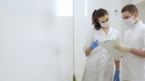 Двоє маскованих лікарів читають медичні документи і обговорюють діагноз пацієнта в сучасній лікарні під час пандемії карантину — стокове відео