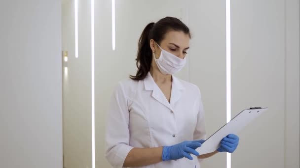 Een vrouwelijke arts in een wit gewaad, masker en handschoenen leest medische dossiers en rapporteert de diagnose aan een camera in een nieuwe kliniek tijdens de pandemie — Stockvideo