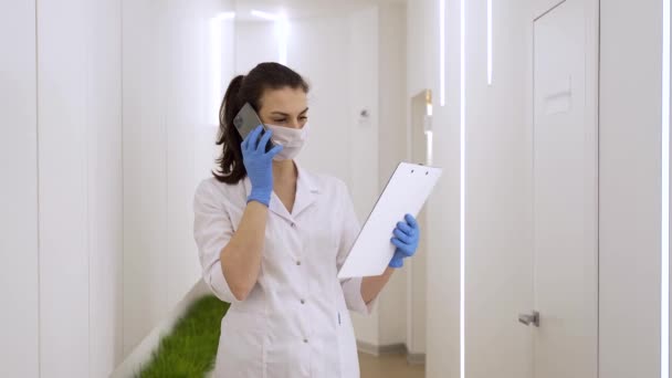 Femme masquée discute de la maladie au téléphone et lit des documents médicaux dans une clinique moderne pendant la pandémie — Video