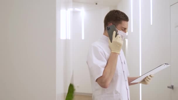 Medico maschile discute la diagnosi del paziente al telefono e cartelle cliniche in una moderna clinica durante la pandemia — Video Stock