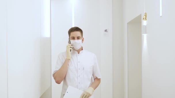 A masked man walks down the corridor at a modern clinic and talks on the phone, holding medical documents in his hand — Stock Video